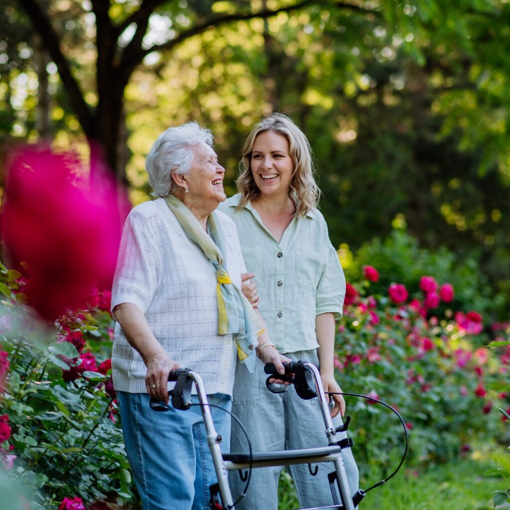 Adult granddaguhter supporting her senior grandmother when taking her for walk with walker in park in summer.