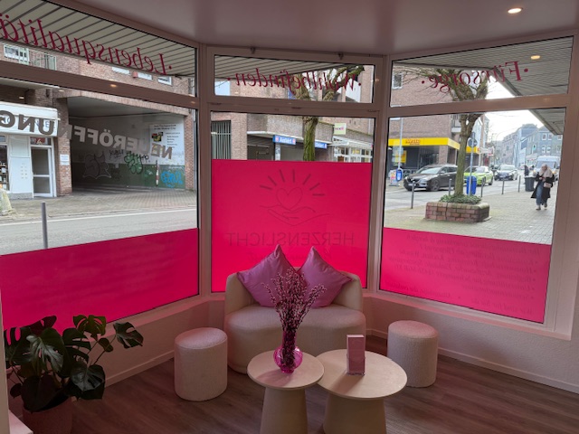 Gemütliche Warteecke mit rosa Dekoration und Pflanzen, breite Fenster mit Straßenausblick.